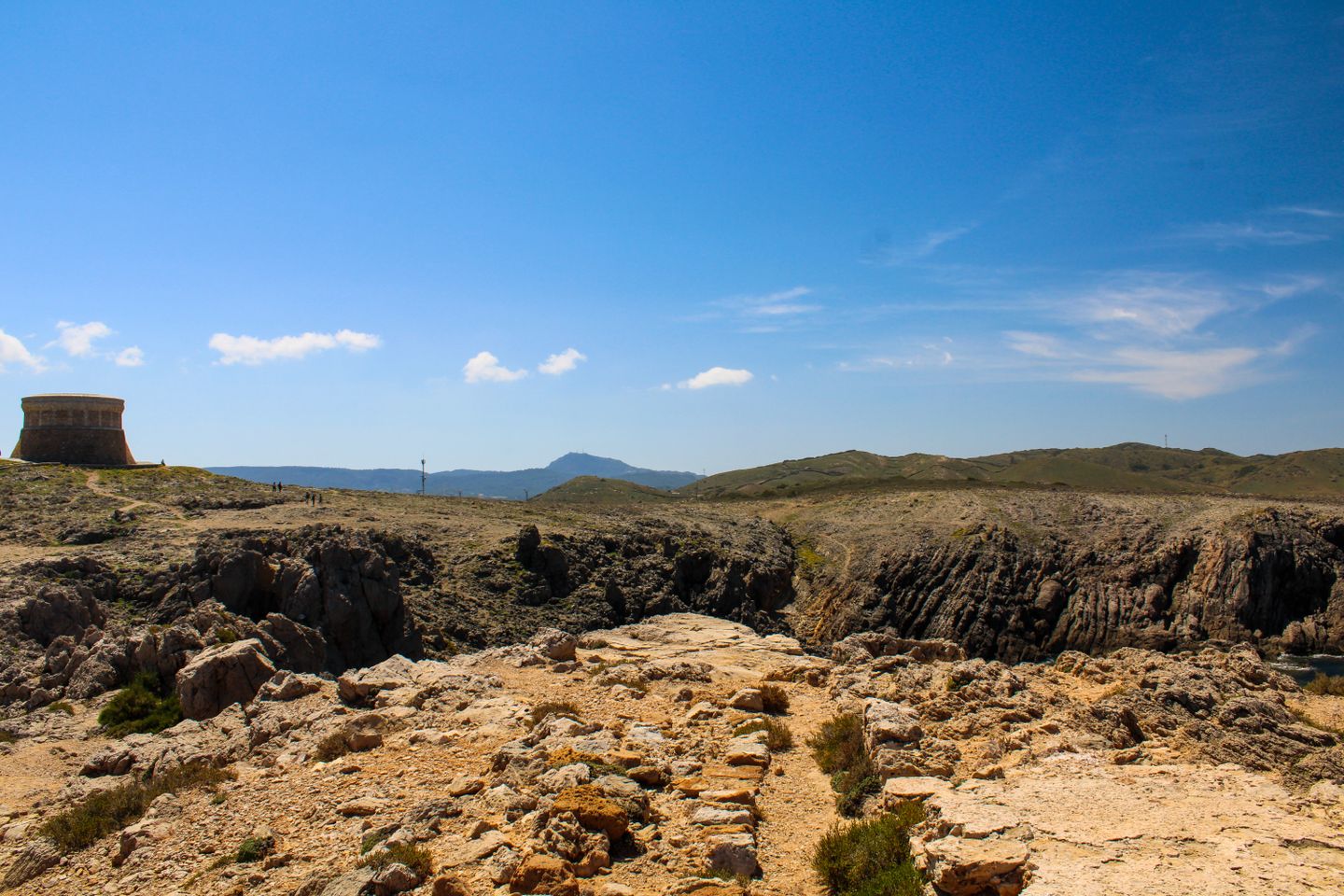 Explora el terreno rocoso y la icónica Torre de Fornelles bajo un cielo azul claro en Menorca, España.