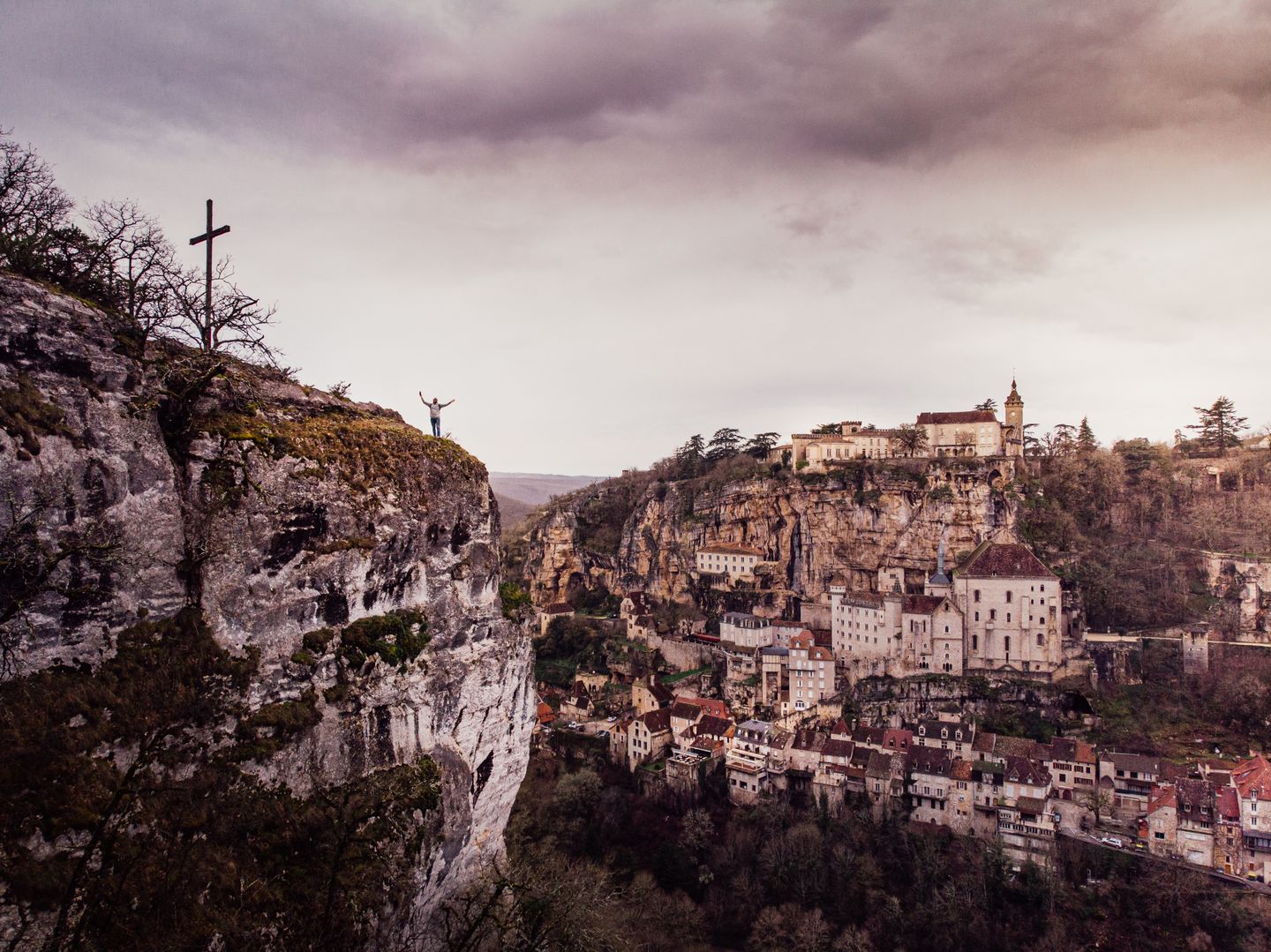 Vista panorámica de la arquitectura medieval de Rocamadour situada en un acantilado, con una persona cerca de una cruz.
