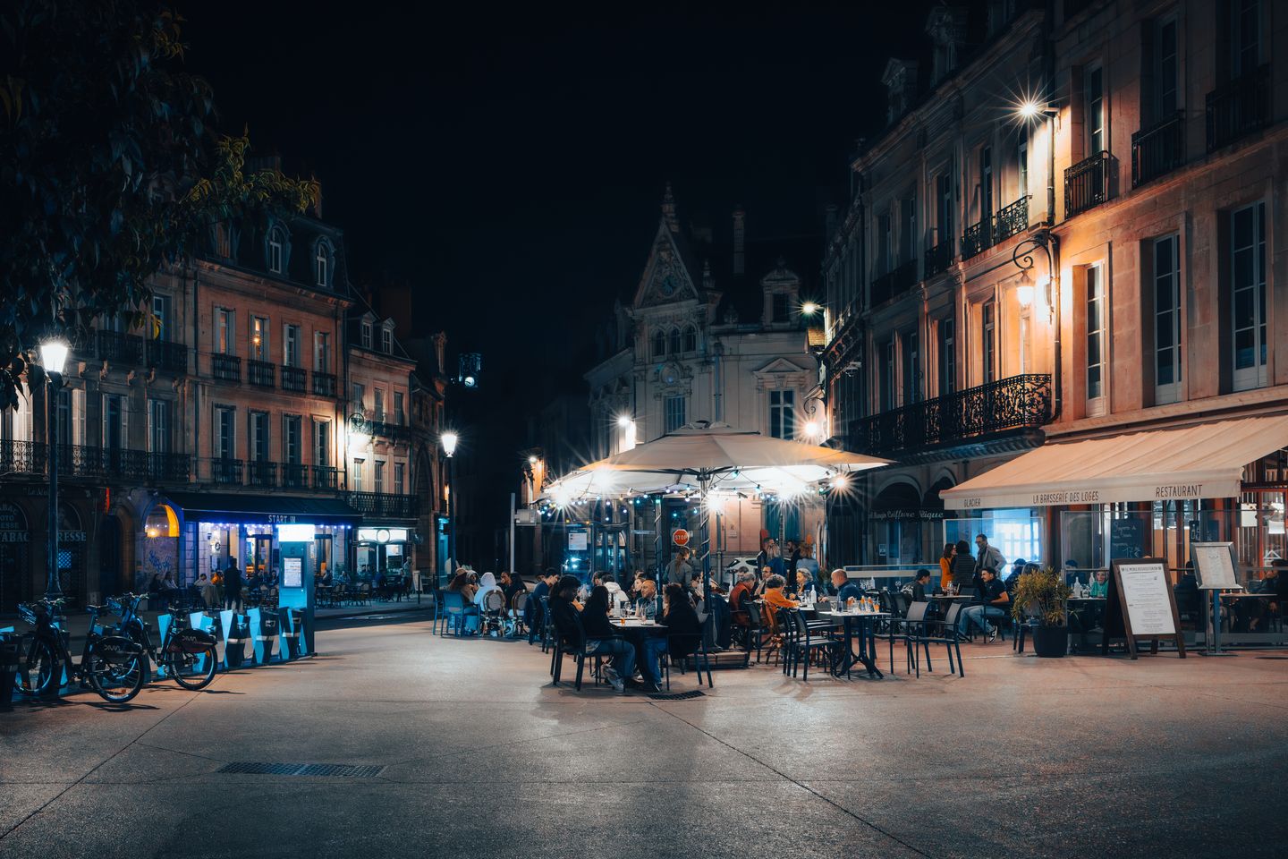 Animada escena nocturna en Dijon, Francia, con una animada cafetería callejera y arquitectura histórica.