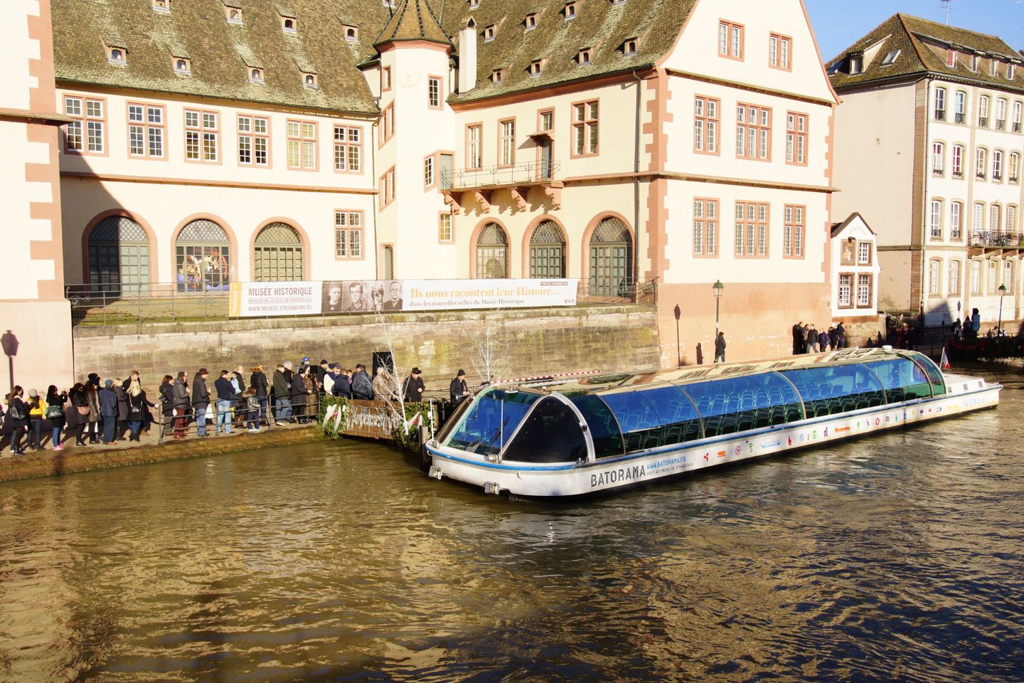 Barco turístico con techo de vidrio navegando junto a la arquitectura histórica de Estrasburgo en un día soleado.