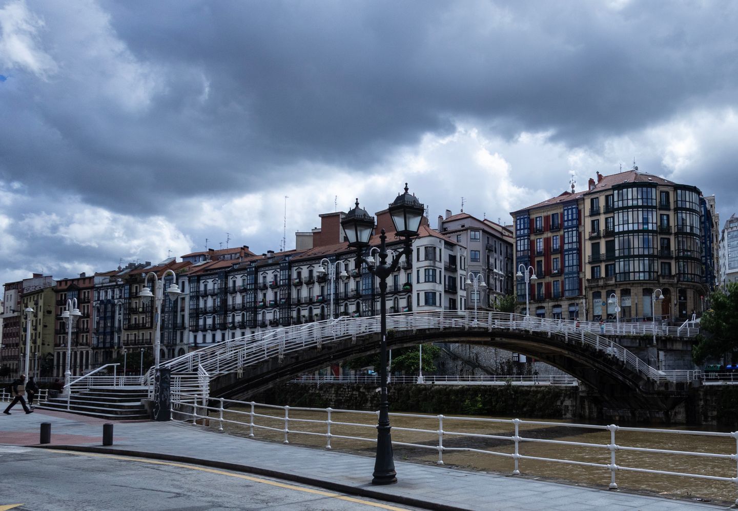 Escena melancólica de Bilbao con un puente histórico y arquitectura tradicional bajo un cielo nublado y dramático.